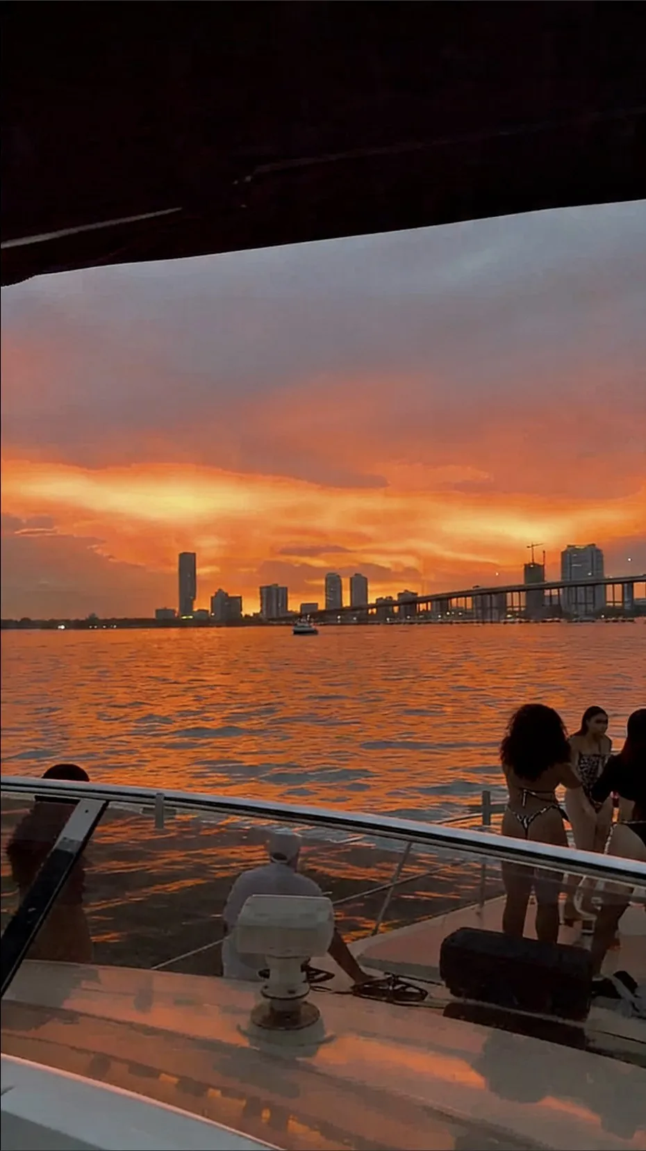 Guests on the foredeck at sunset, the skyline in silhouette across orange water