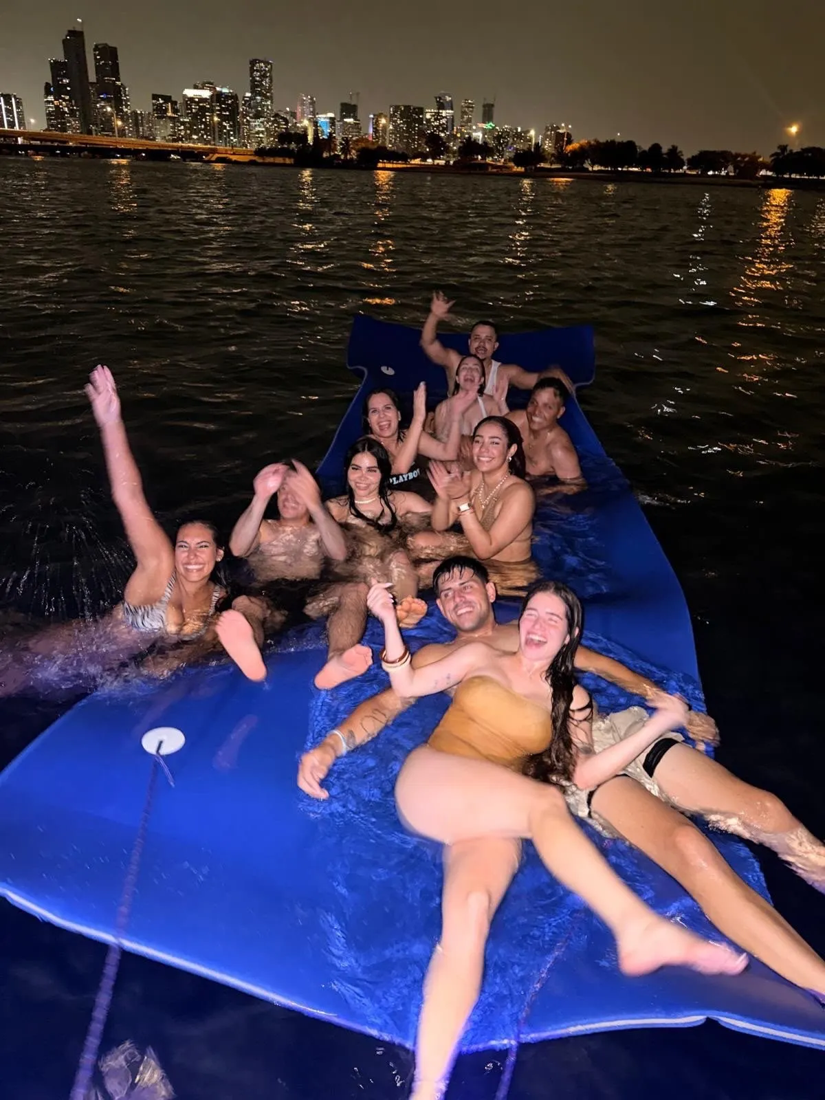A large group lying across the floating water mat at night, the lit Miami skyline behind